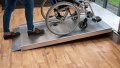 Wide view of a wheelchair ramp without handrails. A wheelchair is being pushed up the ramp which is resting on a step.