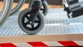 extra wide folding wheelchair ramp with red safety stripes, resting on a step with a manual wheelchair being pushed up. Side view shows the safety tape and grip surface up close