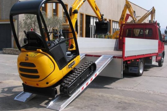 Digger driving up pair of aluminium loading ramps into flat bed van 